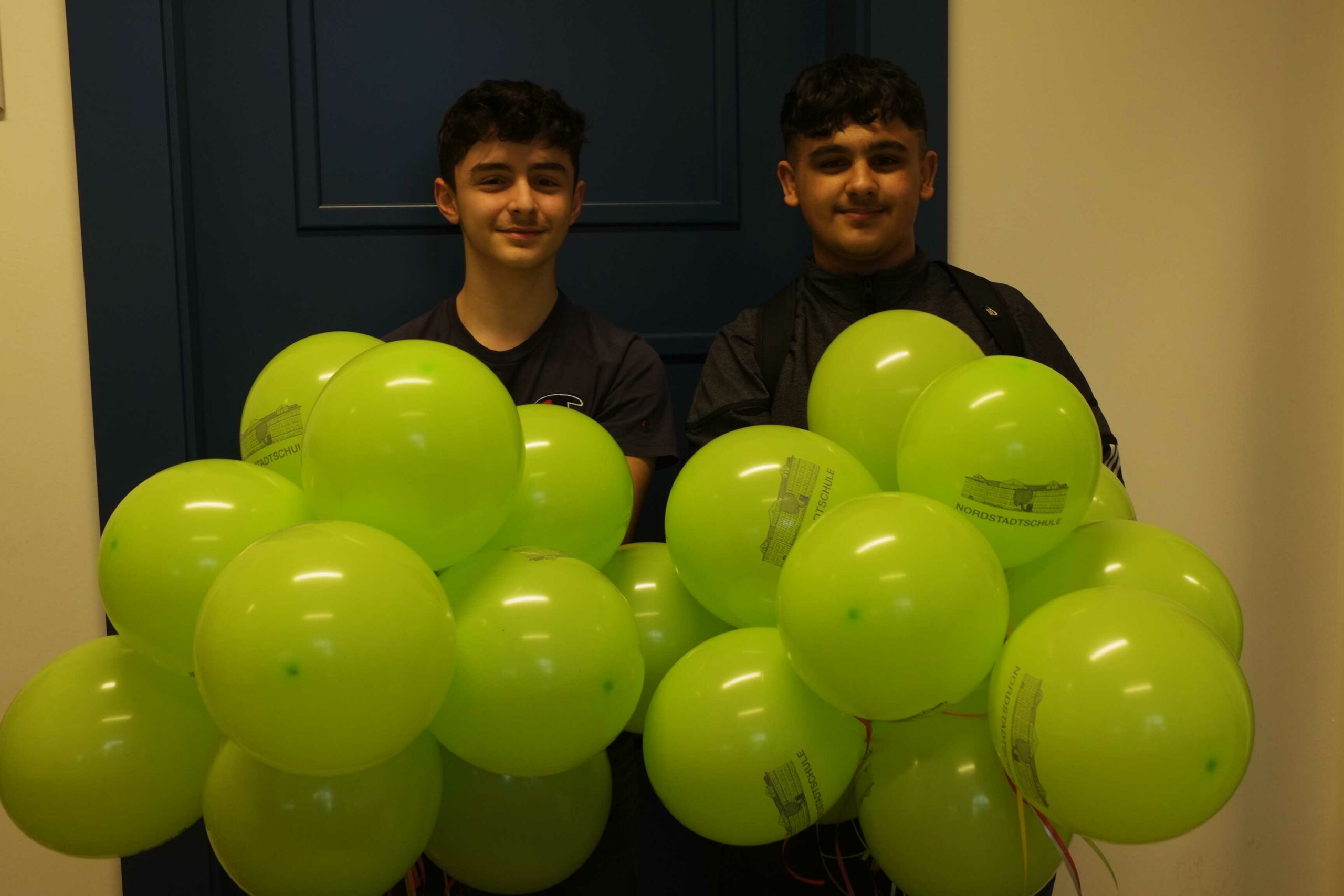 Auf diesem Bild sehen Sie zwei Schüler mit grünen Luftballons in der Hand.