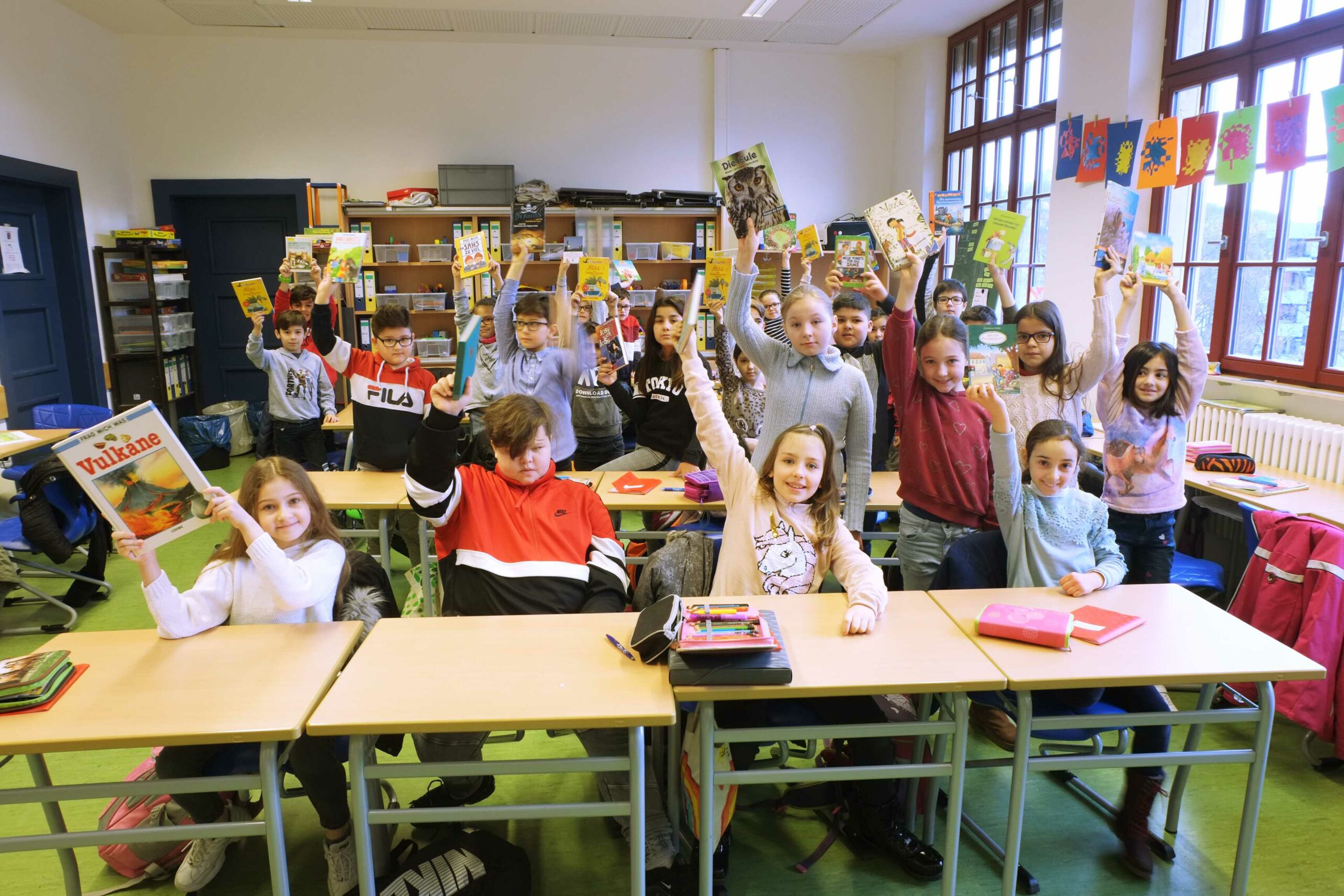 Auf diesem bild sehen Sie Schülerinnen und Schüler in einem Klassenzimmer, welche Bücher in die Luft halten.