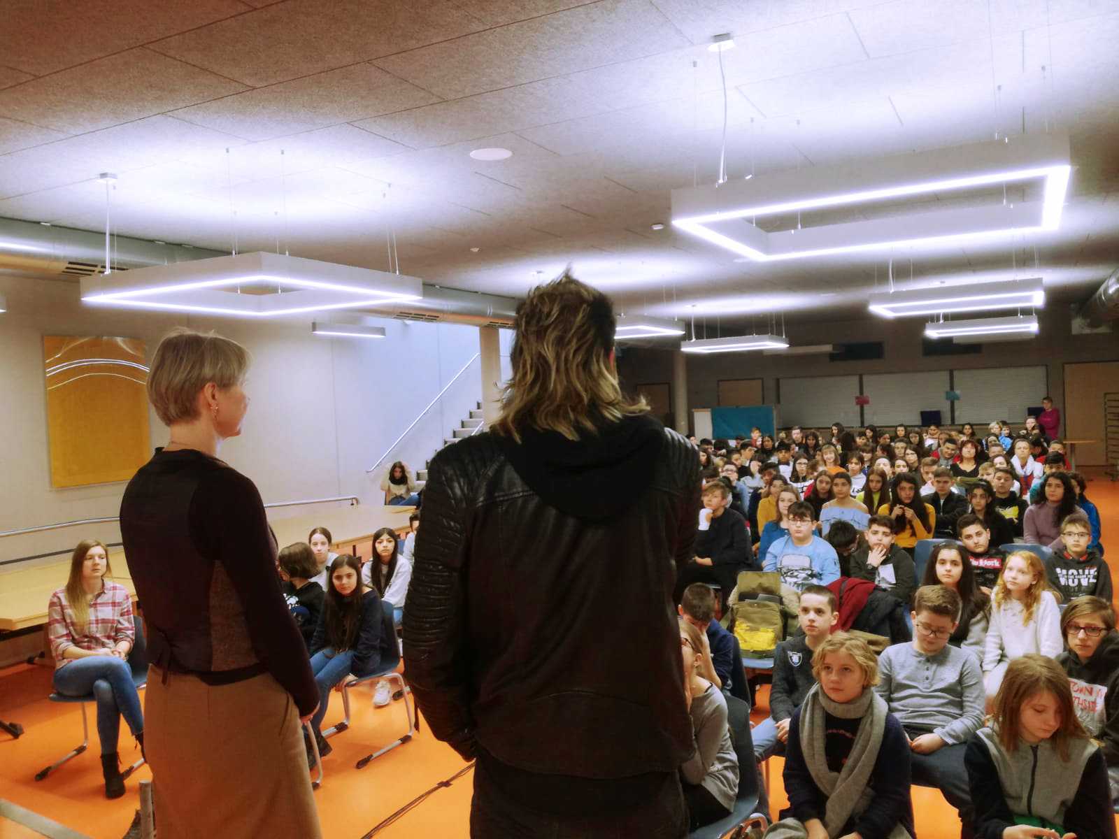 Auf diesem Bild sehen Sie einen Mann und eine Frau auf der Bühne mit einem Mikrofon in der Hand, die zu den Schülern sprechen.