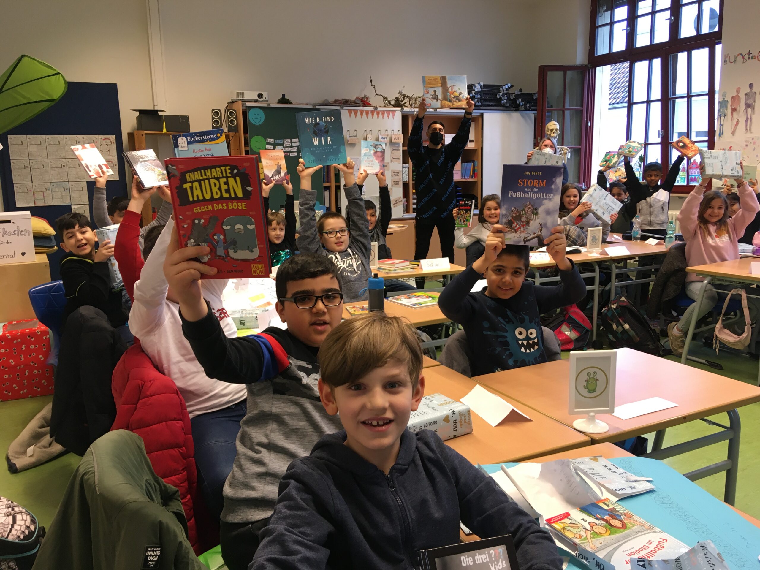 Auf diesem Bild sehen Sie Schülerinnen und Schüler in einem Klassenzimmer, während sie Bücher in die Luft halten.
