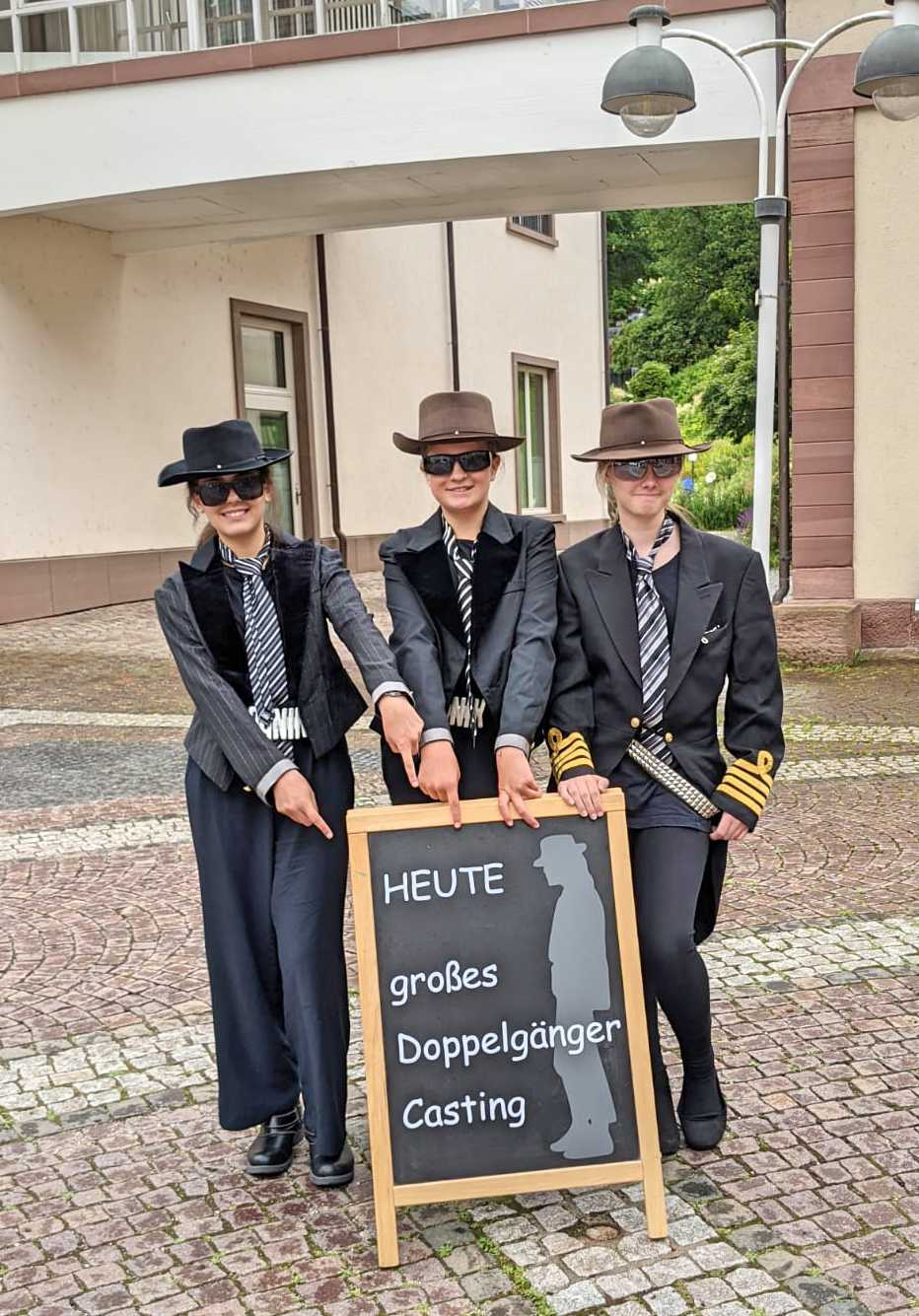 Auf diesem Bild sehen Sie Schülerinnen und Schüler verkleidet als Udo Lindenberg, in schwarz mit Hut und Sonnenbrille in Bad Teinach vor einem Schild mit dem Titel 