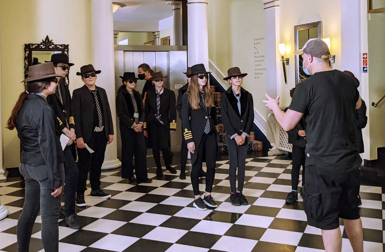 Auf diesem Bild sehen Sie Schülerinnen und Schüler verkleidet als Udo Lindenberg, in schwarz mit Hut und Sonnenbrille in einem Saal.