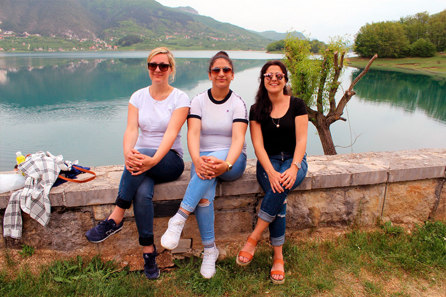 Auf diesem Foto sehen Sie drei Lehrerinnen, die auf einer Mauer sitzen, vor einem See in Bosnien Herzegowina.