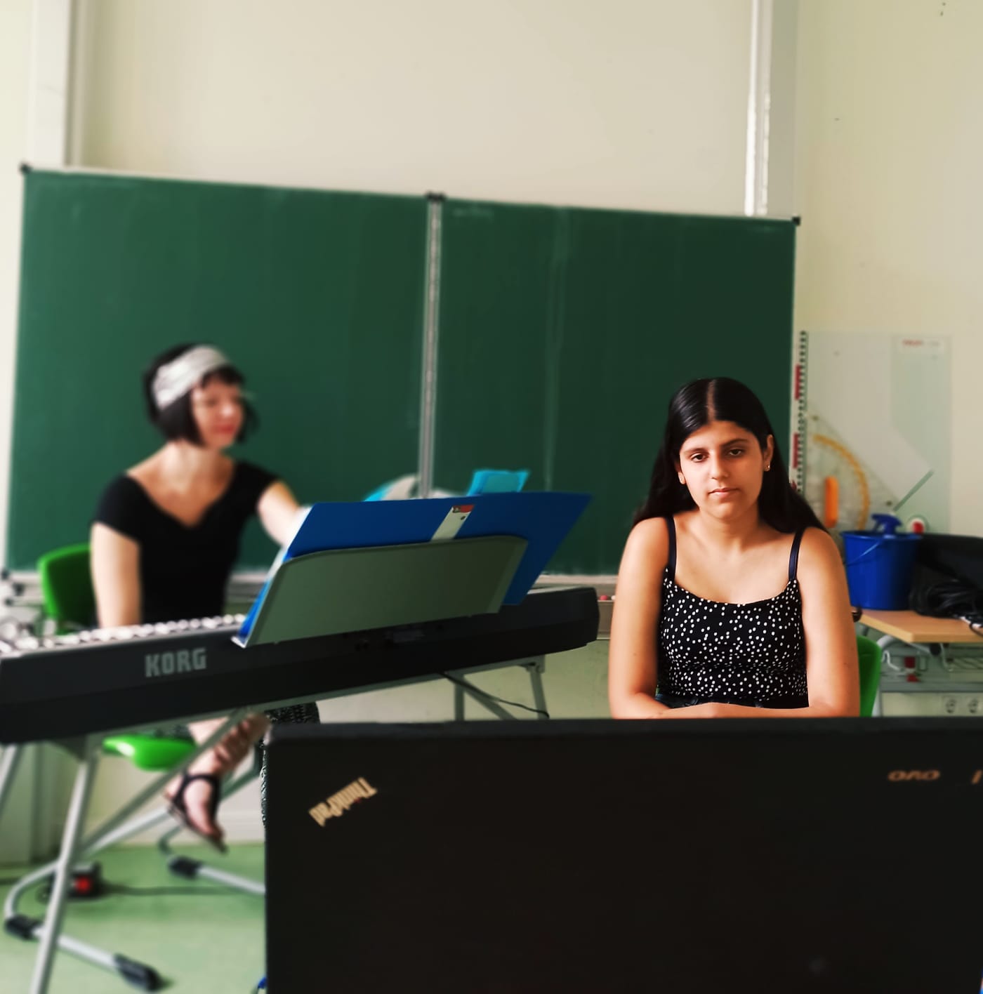 Auf diesem Bild sehen Sie eine Schülerin vor dem Laptop. Hinter ihr sitzt eine Lehrerin am Klavier.