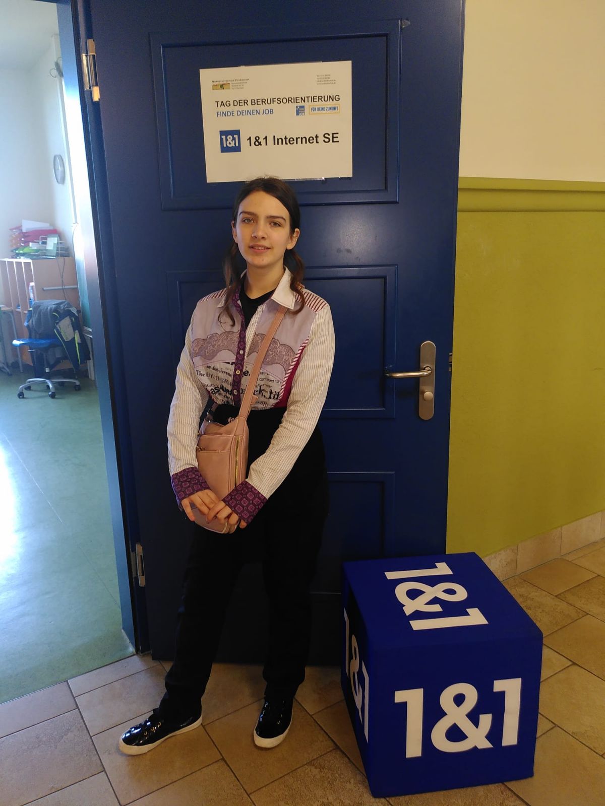 14. Eine Schülerin steht vor einer Tür mit der Aufschrift „der Tag der Berufsorientierung, 1&1 Internet SE“ neben ihr steht ein grober, blauer Würfel mit dem 1&1 Logo auf jeder Seite.